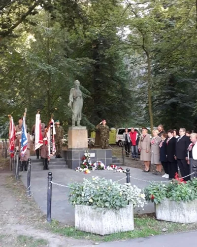 Pietní akt u pomníku vojáka v parku; účastníci stojí v řadě, vojáci salutují, vztyčené prapory a položeny věnce u podstavce