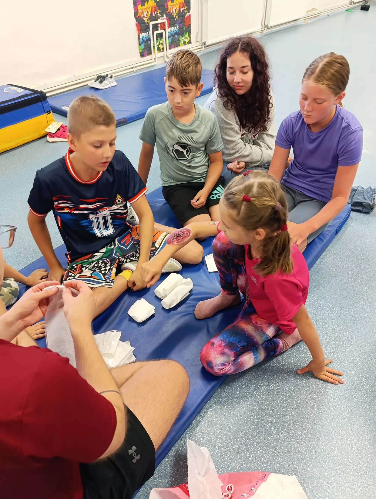 Skupina dětí sedí na modré cvičební podložce, dospělý jim předvádí ošetření rány a techniky obvazování.