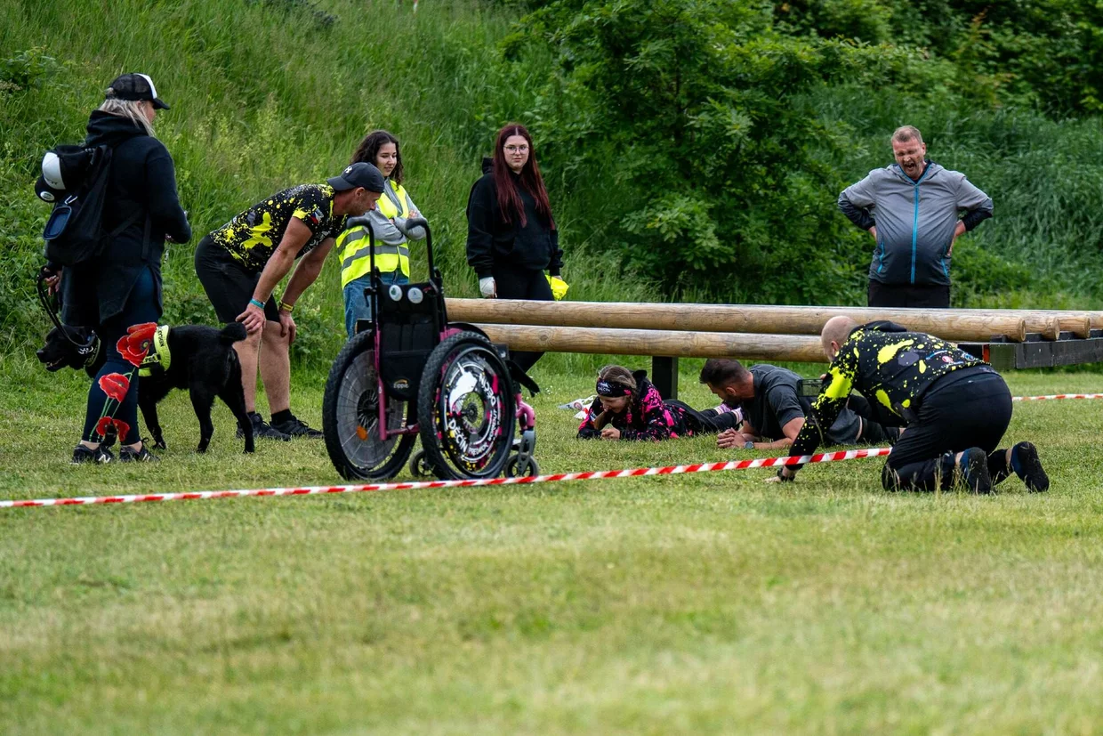 Lidé na venkovním závodě pomáhají účastníkovi na invalidním vozíku přelézt dřevěnou překážku; další soutěžící leží na břiše pod překážkou, poblíž stojí pes a dobrovolníci s reflexními vestami.