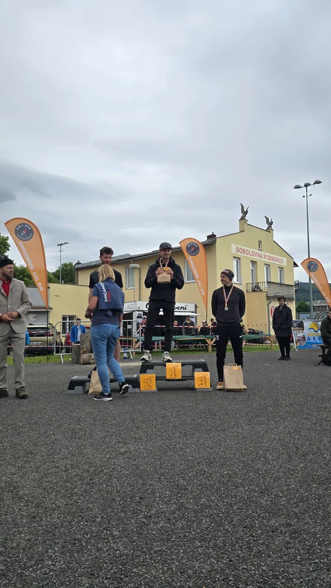 Vyhlášení vítězů venkovního závodu na pódiu před budovou; muži s medailemi a cenami, oranžové praporky s logem, zatažená obloha.