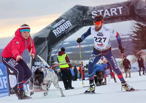 Skijoringový závod: muž na běžkách s číslem 227 tažený psem, vedle něj organizátorka v červené bundě u startovní brány na zasněžené trati.