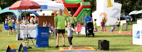 Dítě skáče na měkké podložce za doprovodu dospělého dobrovolníka na rodinné akci v parku; v pozadí nafukovací hrad a stánky.