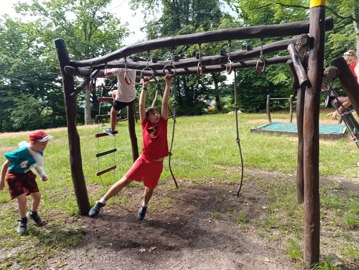 Děti si hrají na dřevěné šplhací konstrukci s kruhy a lany v parku; jedno dítě visí na kruzích, další stojí u vstupu.