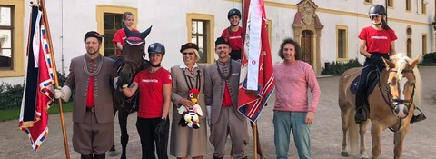 Skupina lidí v historických uniformách a v červených tričkách stojí vedle dvou koní a drží vlajky před barokním zámkem.