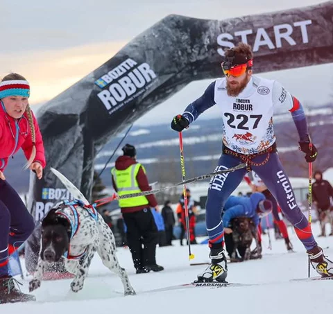 Skijoringový závod: muž na běžkách s číslem 227 tažený psem, vedle něj organizátorka v červené bundě u startovní brány na zasněžené trati.
