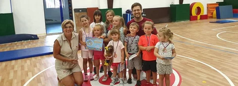 Skupina dětí s medailemi a certifikátem pózuje s dvěma vedoucími v tělocvičně po sportovní aktivitě.