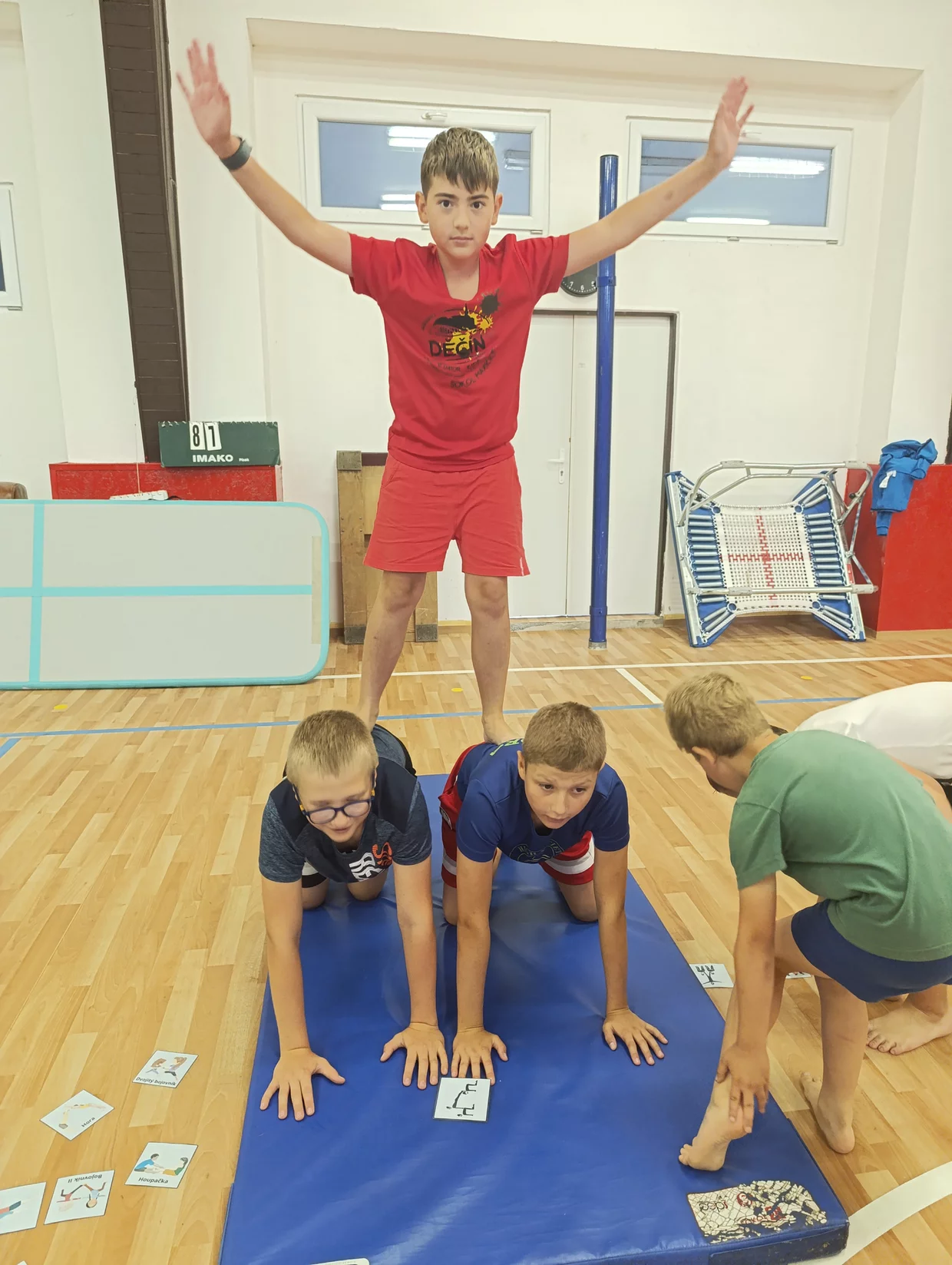 Čtyři děti v tělocvičně tvoří lidskou pyramidu na modré cvičební podložce, jedno dítě stojí nahoře s rozpaženýma rukama a další dítě pomáhá z boku.