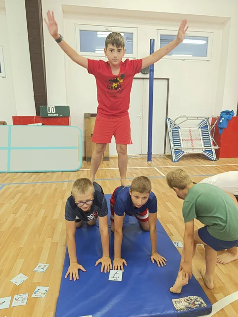 Čtyři děti v tělocvičně tvoří lidskou pyramidu na modré cvičební podložce, jedno dítě stojí nahoře s rozpaženýma rukama a další dítě pomáhá z boku.