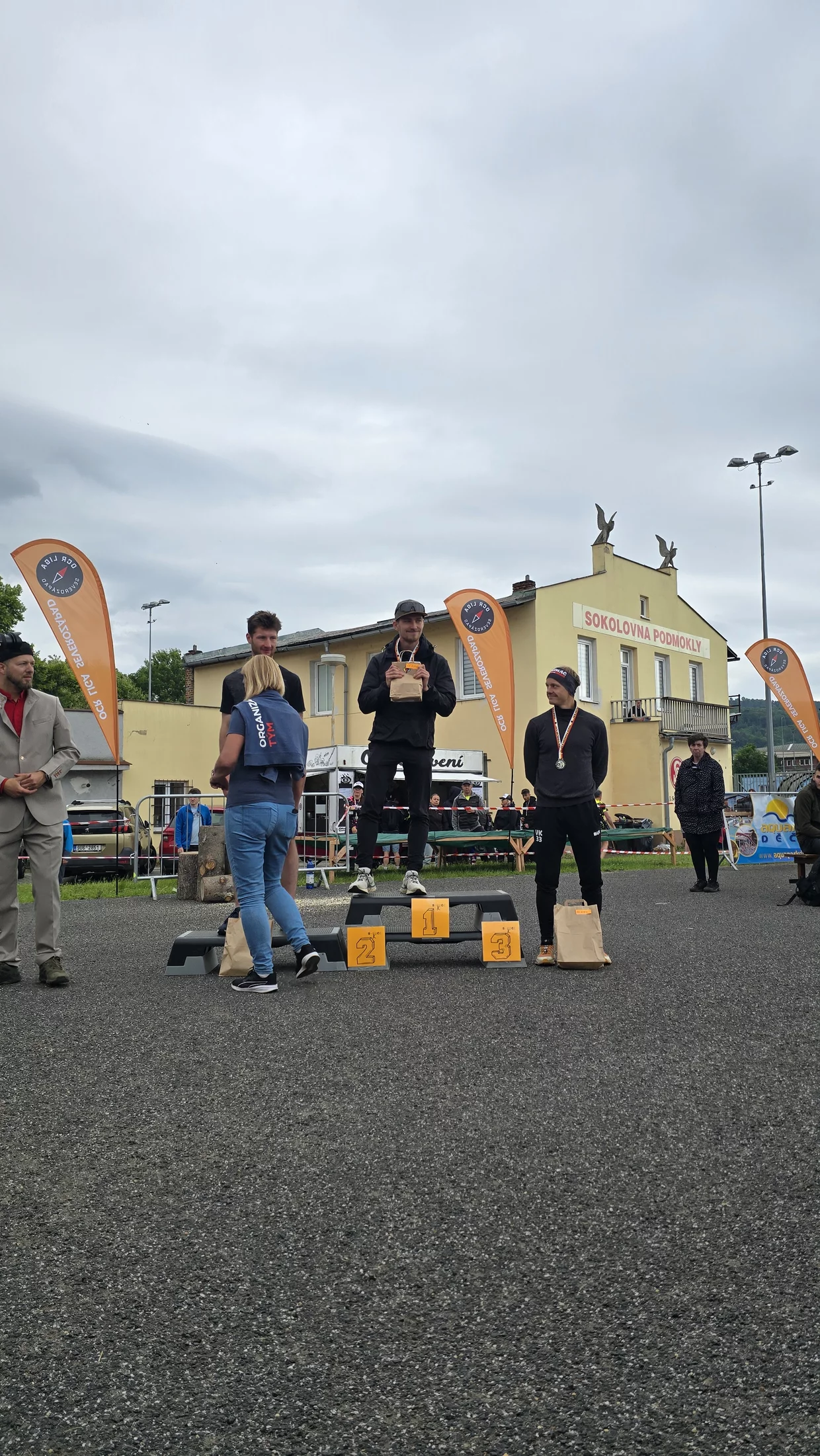 Vyhlášení vítězů venkovního závodu na pódiu před budovou; muži s medailemi a cenami, oranžové praporky s logem, zatažená obloha.