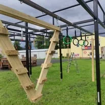 Venkovní překážková dráha s dřevěným V-žebříkem a zelenými gymnastickými kruhy zavěšenými na kovovém rámu, travnaté hřiště a budovy v pozadí.