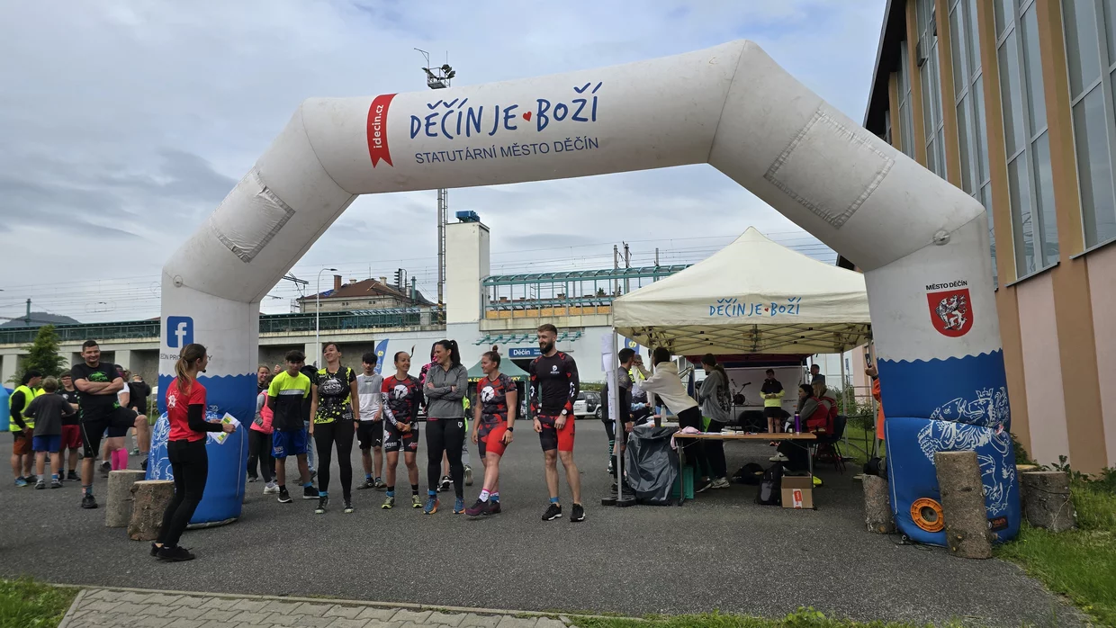 Skupina běžců a organizátorů stojí pod nafukovací bránou s nápisem 'Děčín je boží' vedle registračního stanu u startu závodu