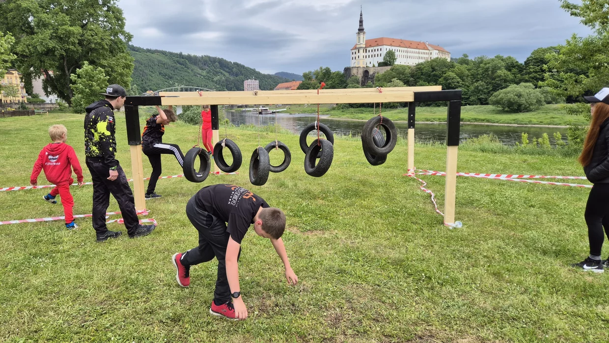 Děti a dospělí překonávají překážkovou dráhu s visícími pneumatikami na trávníku u řeky, v pozadí zámek na návrší.