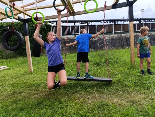 Tři děti si hrají na venkovní překážkové dráze na trávě; dívka visí na kruzích, jeden chlapec stojí na houpačce a další chlapec stojí vpravo.