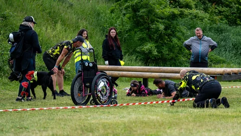 Lidé na venkovním závodě pomáhají účastníkovi na invalidním vozíku přelézt dřevěnou překážku; další soutěžící leží na břiše pod překážkou, poblíž stojí pes a dobrovolníci s reflexními vestami.