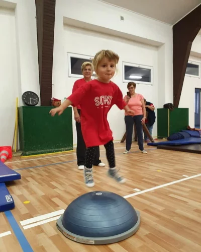 Dítě skáče přes půlkouli bosu v tělocvičně během cvičení, v pozadí dospělí sledují.