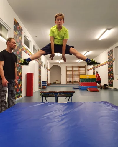 Chlapec v žlutozeleném tričku skáče roznožku ve vzduchu při přeskoku přes trampolínu v tělocvičně, vedle pozoruje trenér.