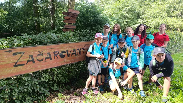 Skupina dětí v modrých tričkách a s batohy stojí u dřevěného nápisu "Začarovaný" v zeleném lesním parku.